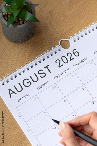 Hand holding a pen on August 2026 calendar. Top view.