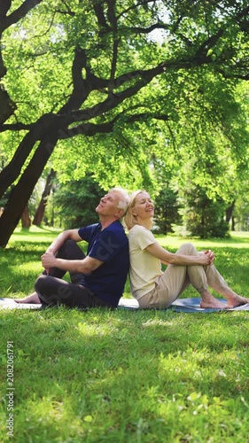 Smiling senior couple sitting back to back on yoga mats under large tree in sunny park. Mature man and woman relaxing after outdoor workout