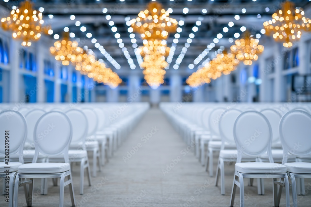 Obraz premium Rows of empty white chairs in a spacious hall illuminated by ornate blurred chandeliers, ready for a grand event.