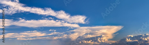 A diagonal dynamic formation of broad evening atmospheric front. Yellow, tawny pastel clouds hang solemnly over horizon against blue sky. The differential volume of a rain cloud. Copy space for text.