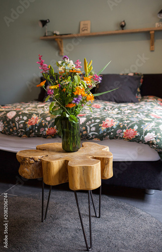 Colorful wildflower bouquet in tall green glass vase on wooden stump side table with floral bedding background . High quality photo