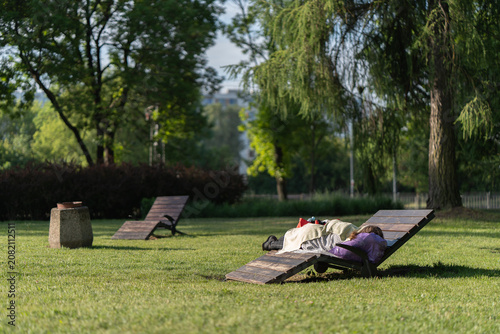 Foto Homeless woman sleeps on wooden lounge chair in a public park representing poverty, social issues, vagrancy and loneliness while resting near belongings in a summer day