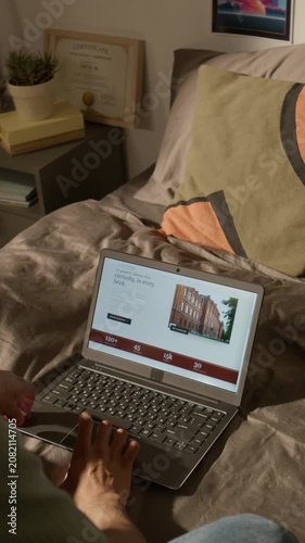 Vertical video of hand of unrecognizable young student lying in bed, scrolling through college website on laptop. Clip is ideal for university enrollment ads and higher education promotional videos