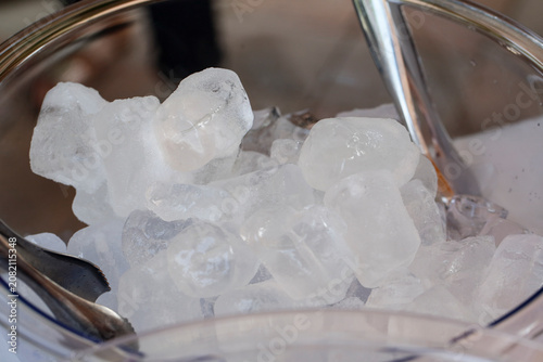 Transparent ice cubes in a clear glass bowl with metal tongs. The scene captures a fresh and cool ambiance suitable for beverages and summer settings.