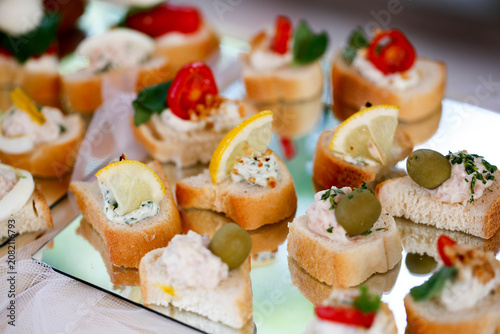 A variety of elegant canapes featuring bread, cherry tomatoes, olives, lemon wedges, and creamy toppings placed on a reflective surface for vibrant presentation.