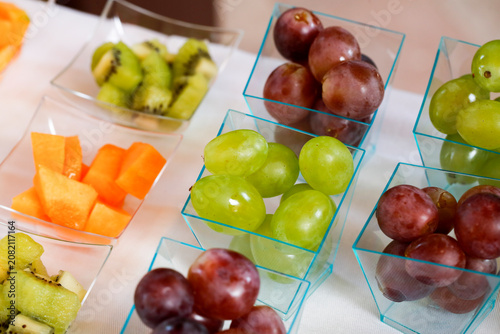 Assorted fruits displayed in transparent bowls on a white surface, featuring grapes, kiwi, and melon cubes with vibrant and inviting colors.