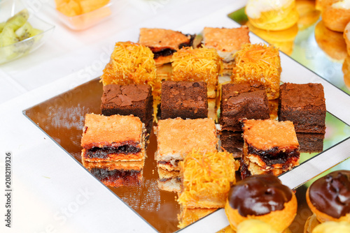 A variety of desserts including chocolate brownies, flaky fruit bars, and cream puffs displayed on a shiny gold surface with a neat presentation.