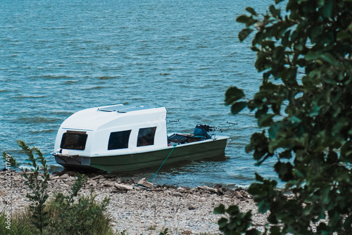 Small cabin boat moored beside rocky lake shore with rippled water and green foliage, concept of boating, fishing, lakeside travel and quiet outdoor recreation.