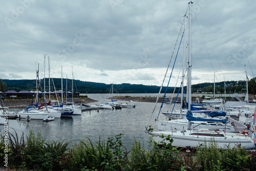 Sailboats moored in a lakeside marina under dramatic cloudy sky with forested hills, calm water and quiet outdoor travel atmosphere.