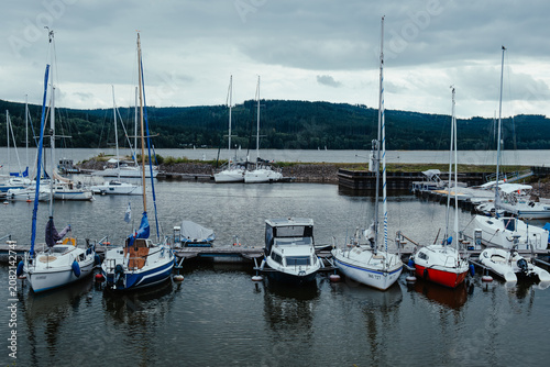 Sailboats and small boats moored in a quiet lake marina with cloudy sky, calm water, reflections and forested hills in the background.