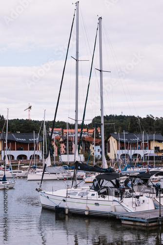 White sailboat moored at dock in a lakeside marina with other boats, calm water, waterfront houses and cloudy sky in the background.