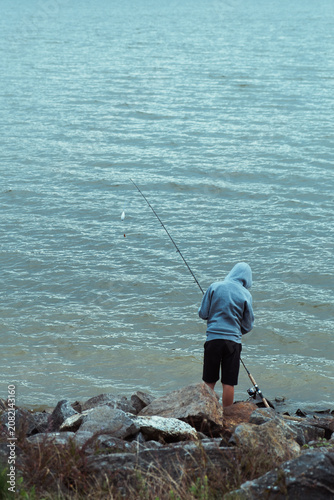 person in hoodie fishing with rod from rocky lakeshore near rippled water, concept of outdoor recreation, solitude, leisure and quiet nature escape.