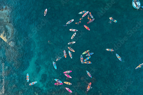 Many people are paddleboarding and swimming in vibrant turquoise ocean waters. Numerous colorful stand up paddleboards dot the clear sea on a bright day