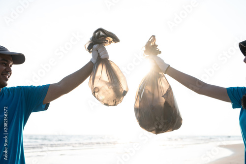 Two happy volunteers smiling and talking while collecting plastic bottles into a trash bag on the beach, enjoying their environmental community service together.