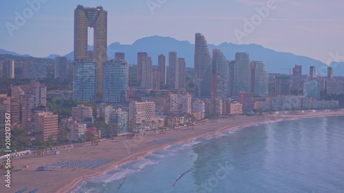 People stroll along the beach as the sun sets behind the buildings. The city skyline stands tall against the mountains in the distance. Waves gently lap at the shore.