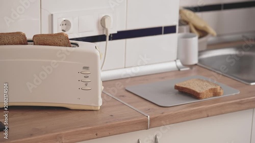 In a kitchen, a toaster warms slices of bread on a counter. The kitchen has a sink and other kitchen tools. The setting is bright in the morning hours.