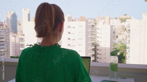A person sits at a table on a balcony and uses a laptop. The city skyline and plants are visible in the background. It is a bright day with clear skies.