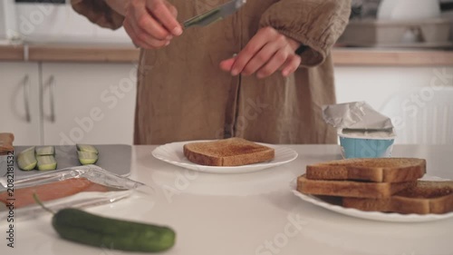 A person prepares toast in a kitchen. They take slices of bread, spread cream cheese on top, and add cucumber. This takes place in a home kitchen.