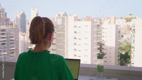 A woman sits on a balcony and uses her laptop. She looks out at the city buildings during the day, enjoying the view. A small plant sits beside her on the table.