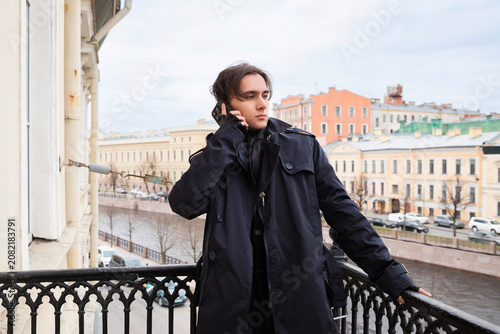 Young stylish man talking on mobile phone on balcony. Fashionable guy wearing black trench coat outside during city day. Communication and business lifestyle concept in urban area.