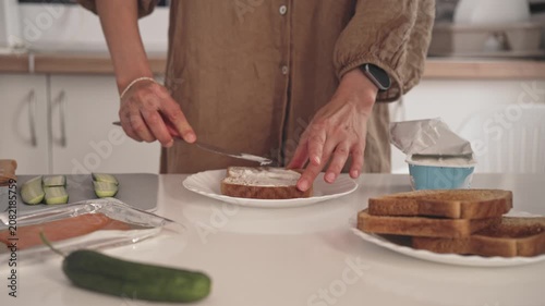 Hands spread cream cheese on a slice of toast in the kitchen. Cucumber slices are placed alongside. Bread is prepared for an easy meal.