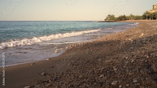 Waves come and go on the sandy beach as the sun hovers low in the sky. Water splashes softly against rocks while palm trees stand in the background.