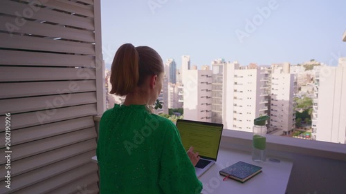 A person is standing on a balcony. They are using a laptop while looking at the city. Buildings are visible in the background. The sky is clear and blue.