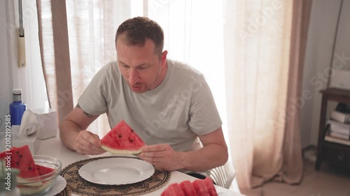 A person sits at a table in a kitchen. He picks up pieces of watermelon and eats them. Sunlight comes into the room through the window. Friends sit nearby and enjoy the moment.