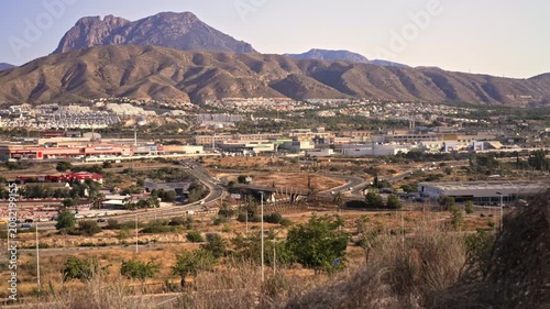 A view shows mountains and a city landscape from a high point. The sun shines bright, and the skies are clear. The city is busy with roads and buildings visible.