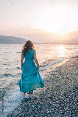 Young woman in a light dress stands by a waterfront railing, enjoying a peaceful sunset over the sea.