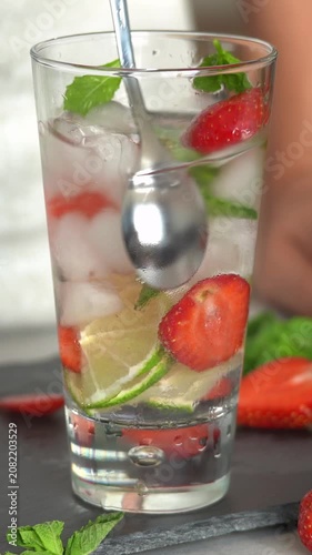 Hands stir fresh strawberry Mojito cocktail with lime strawberry mint ice and soda in glass. Cocktail preparation. Vertical shot