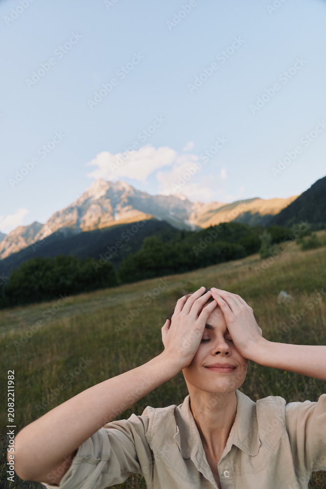 Fototapeta premium A woman outdoors in a mountain meadow holds her head with both hands, smiling softly. A moment of relief and self care in nature, capturing a lifestyle scene of calm, balance, and personal reflection.