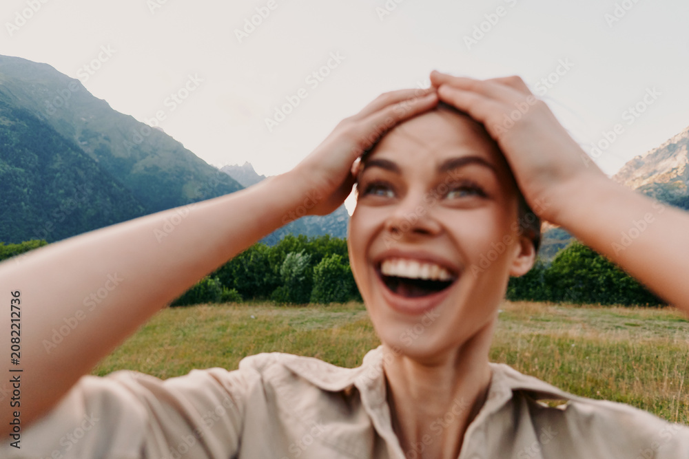 Fototapeta premium Young woman outdoors smiles broadly, hands on head in a sunny meadow with mountains behind, natural light, fresh air, carefree vibe.