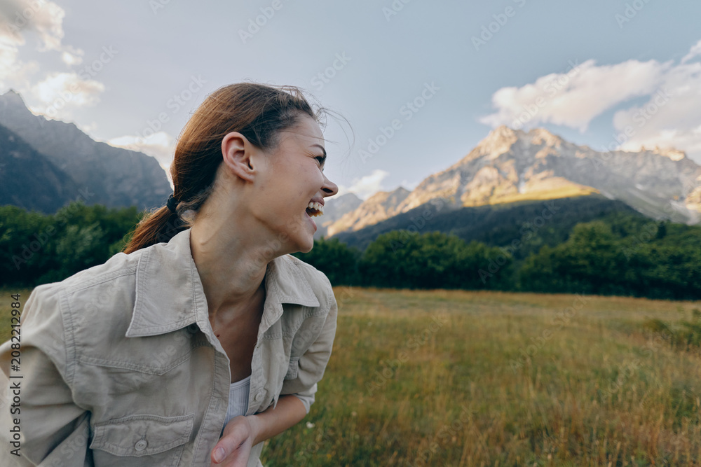 Fototapeta premium Young woman laughs in an open meadow with distant mountains behind, enjoying a sunny day, light breeze, and relaxed posture in natural surroundings