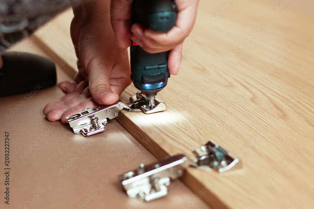 Fototapeta premium Close-up of a person using a power drill to attach metal hinges to a wooden cabinet door during home assembly, with a focus on the tools and materials involved