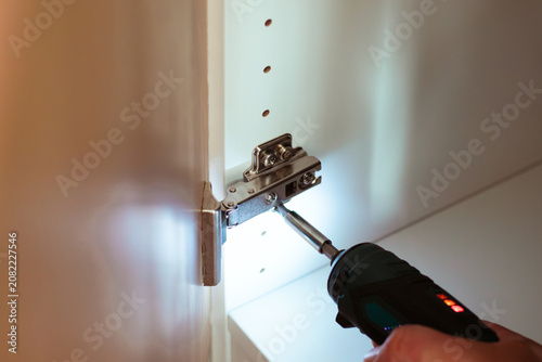 Fotografía Hand holding electric screwdriver tightening cabinet hinge inside a home, illumi