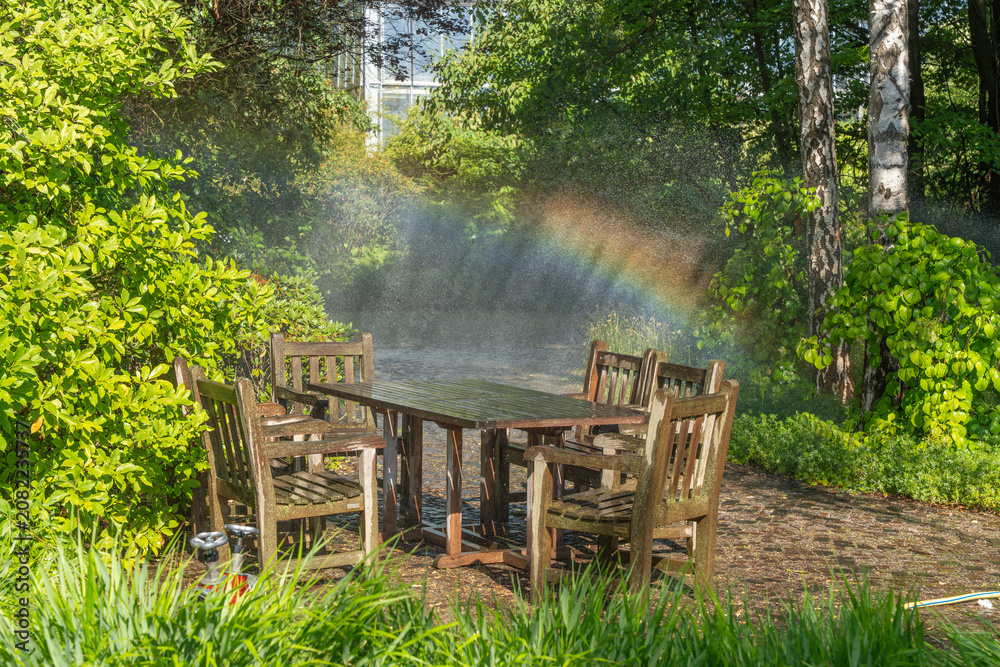 Naklejka premium A wooden patio set sits beneath cooling water mist. A colorful rainbow appears among lush trees and shrubs.