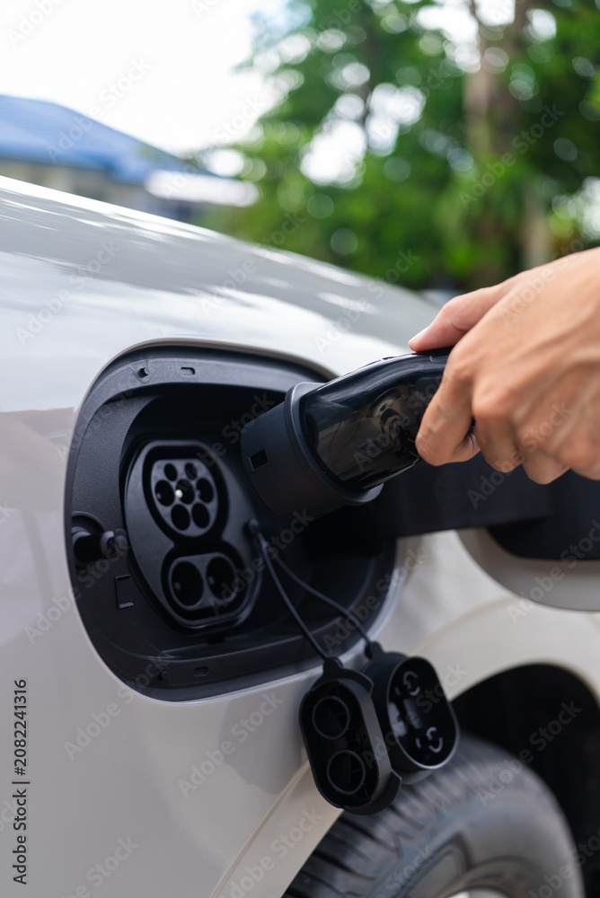 Fototapeta premium Side view of an Asian woman's hand refuel an electric vehicle with a charger plug. Concept of alternative power infrastructure green tech and sustainability. Vertical Shot