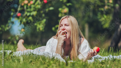 Εκτύπωση καμβά Blond woman reclining on grass blanket, biting crisp apple among fruit trees, ba