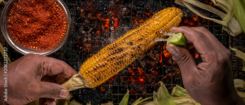 Top-down close-up of hands squeezing lime on roasted Pakistani street corn bhutta — masala spices, glowing charcoal grill, steaming monsoon street food flat lay