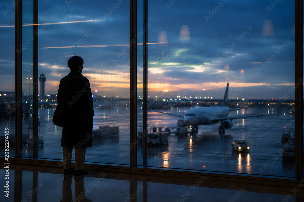 Fototapeta premium Traveler standing by a large airport window watching an aircraft on a rainy evening runway, symbolizing travel, departure, anticipation, and journey.