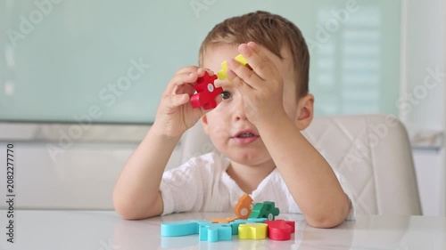 Little child boy plays with colorful plastic building blocks, holds red and yellow pieces while seated at white table. Childhood, early education and children development concept. Slow motion footage