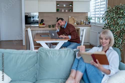Fotografie Man distracted working from home while woman reading on couch