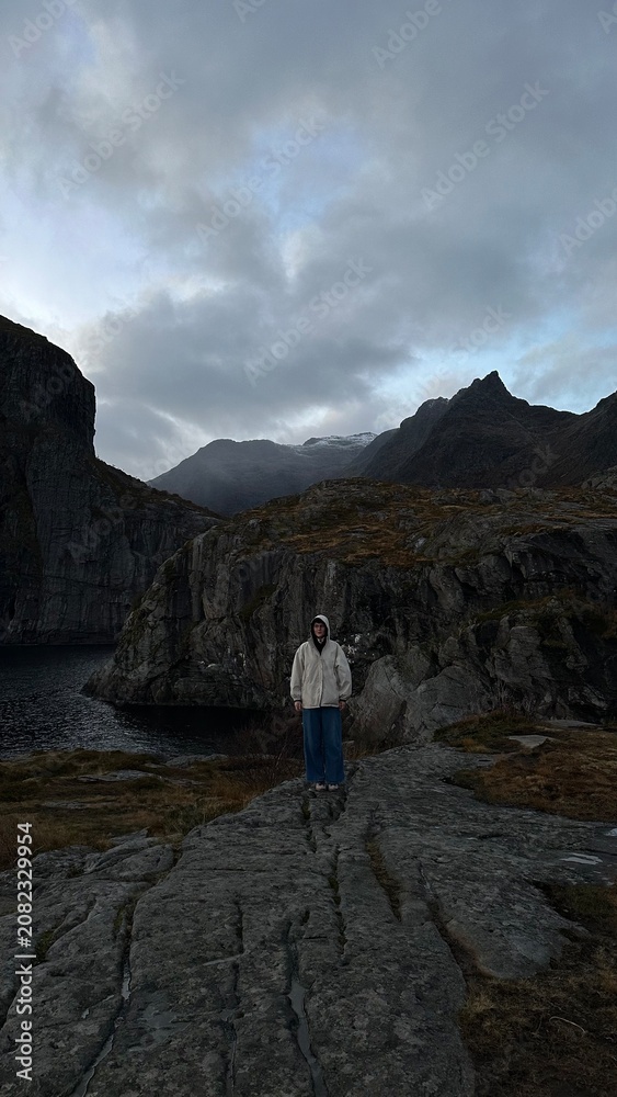 Fototapeta premium Female traveler standing on rocky cliffs in the Lofoten Islands, Norway. Dramatic mountain landscape under cloudy skies, ideal for travel and adventure concepts.