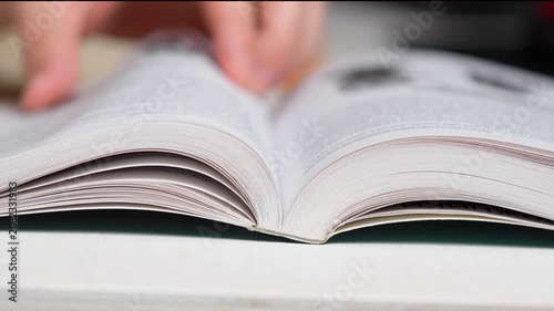 Close-up of a person's hands turning pages of an open book on a desk. Selective focus on open textbook pages, reading, studying, and education concept.