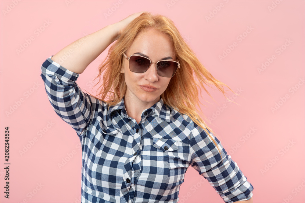 Fototapeta premium Blonde woman posing playfully with her hand in vibrant hair, wearing a checkered plaid shirt and modern sunglasses against a minimal pink background, expressing casual style