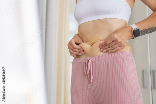 Obraz na plátne Woman standing at home pinching her abdominal fat and love handles with both han