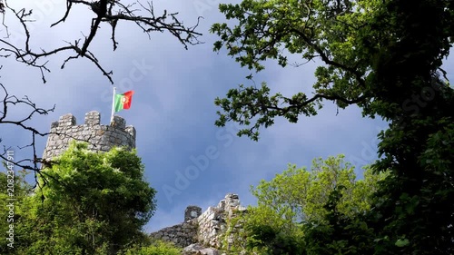low angle view of a medieval stone castle tower with the national flag of portugal waving proudly against a dynamic sky surrounded by green trees