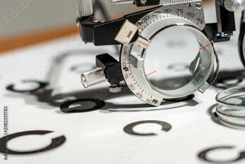 Close-up of an eye measurement device with a circular dial. Dial shows measurement units and a rotating lens. Chart displays letter symbols for vision testing