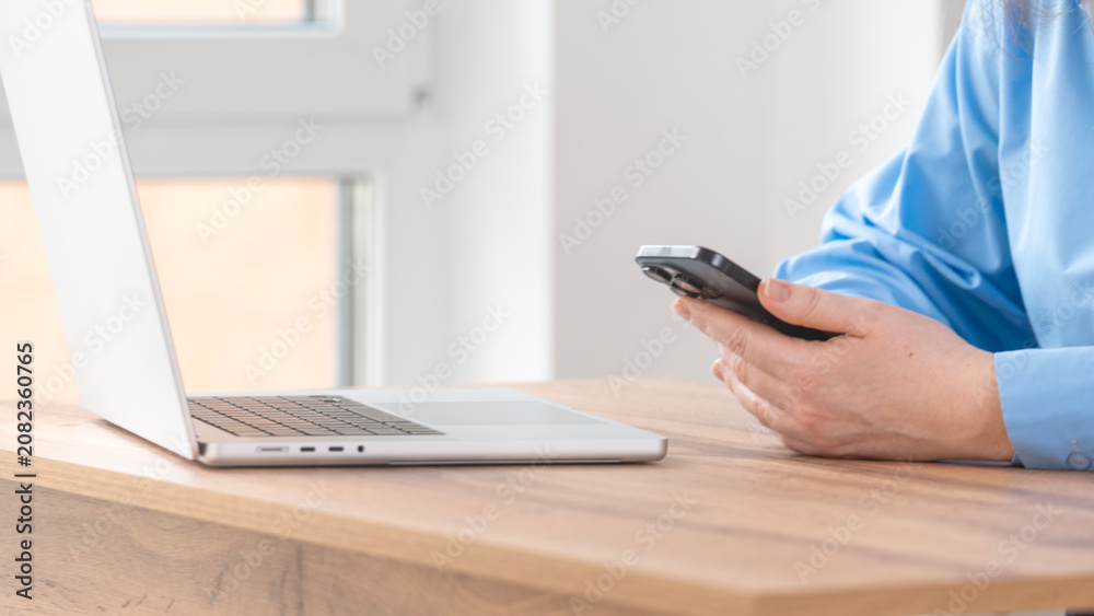 Fototapeta premium Close-up of person's hands holding a smartphone. Silver laptop rests on wooden desk surface. Light streams through window in background. Neutral setting captures modern work environment